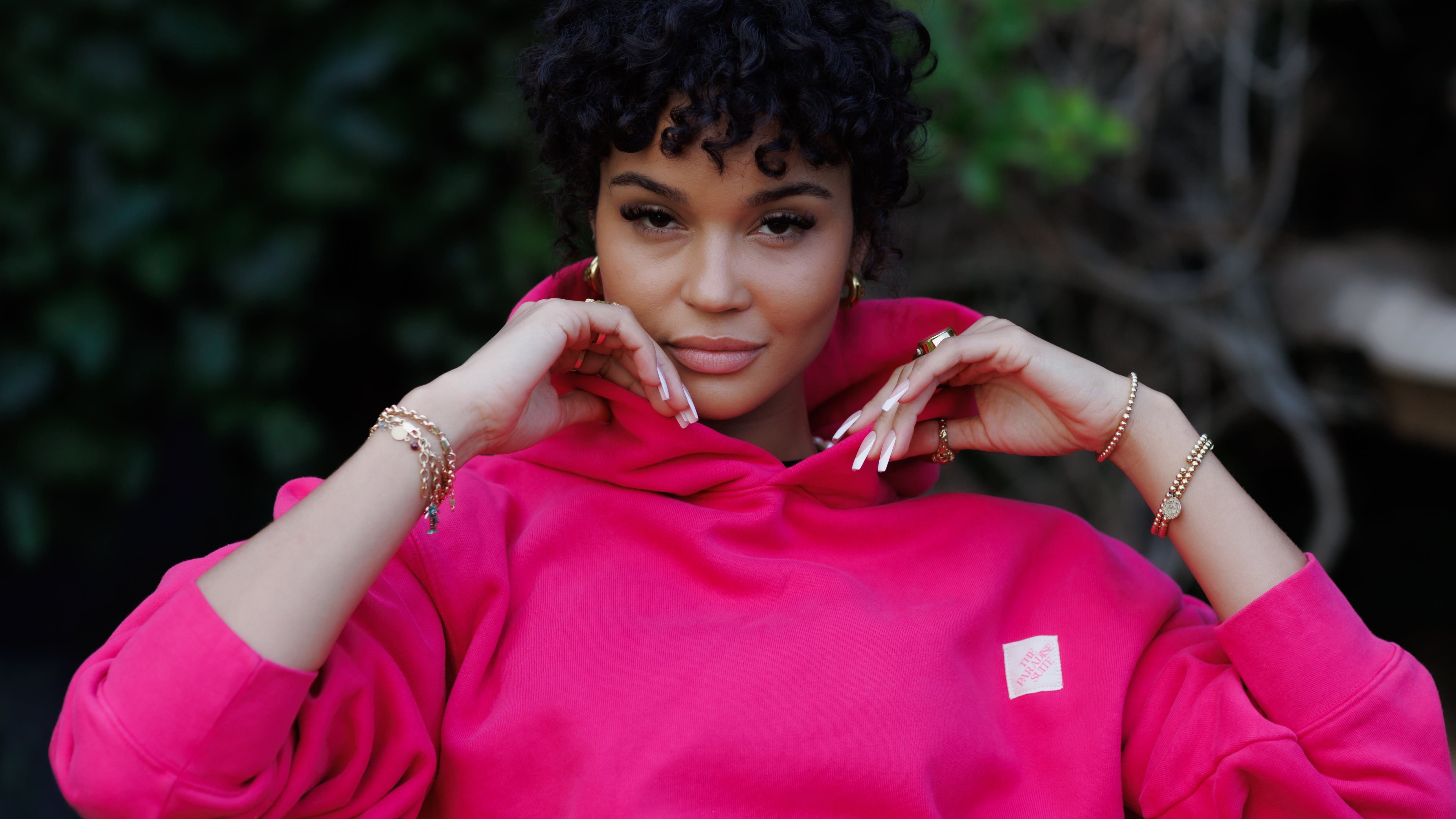 Woman wearing a bright pink hoodie with a small logo, sitting outdoors.